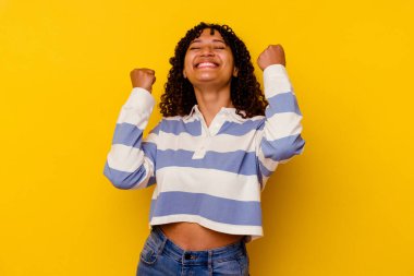 Sarı arka planda izole edilmiş genç melez kadın zaferi, tutkuyu ve coşkuyu kutluyor, mutlu bir ifade..