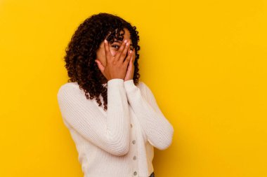 Sarı arka planda izole edilmiş genç melez kadın parmaklarının arasından korkmuş ve gergin bir şekilde göz kırp..