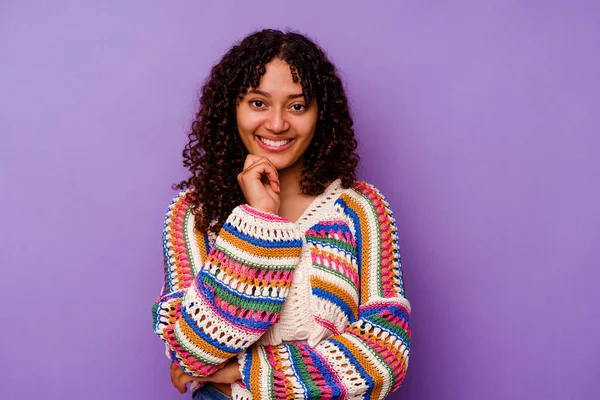 Mor arka planda izole edilmiş genç bir ırk kadını mutlu ve kendinden emin gülümsüyor, elleriyle çenesine dokunuyor..