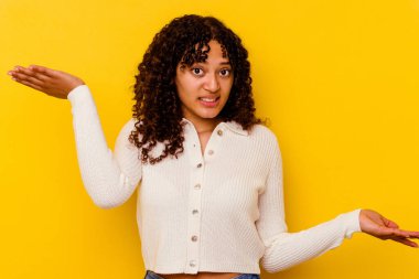 Sarı arka planda izole edilmiş genç melez bir kadın. Kafası karışmış ve omuz silkiyor. Kopya alanını tutmak için..