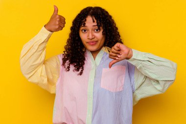 Sarı arka planda izole edilmiş genç, melez bir kadın baş parmağını kaldırıyor, zor seçim konsepti.