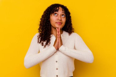 Sarı arka planda izole edilmiş genç melez bir kadın. El ele tutuşup dua ediyor. Kendine güveniyor..
