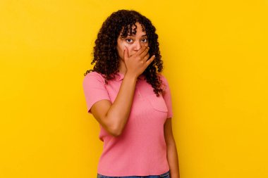 Sarı arka planda izole edilmiş genç melez kadın elleri endişeli bir şekilde ağzı örtüyor..