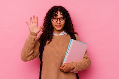 Genç öğrenci melez kadın pembe arka planda izole edilmiş beş numarayı parmaklarıyla gülümsüyor..
