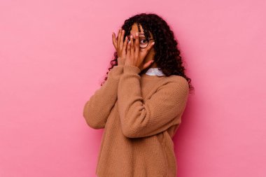 Pembe arka planda izole edilmiş genç melez kadın parmaklarının arasından korkmuş ve gergin bir şekilde göz kırpıyor..