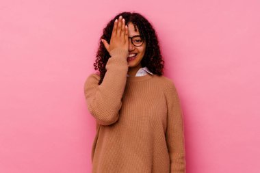 Pembe arka planda izole edilmiş genç melez kadın yüzünün yarısını avuç içiyle kaplayarak eğleniyor..