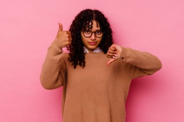 Pembe arka planda soyutlanmış genç melez bir kadın. Baş parmağını kaldırıyor ve onaylıyor. Zor bir seçim.