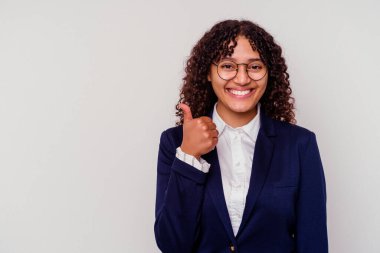 Beyaz arka planda izole edilmiş genç iş kadını gülümsüyor ve başparmağını kaldırıyor.