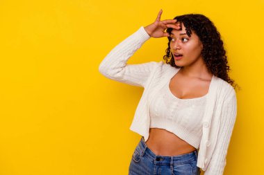 Genç melez kadın, sarı arka planda izole edilmiş. Uzağa bakıyor, elini alnına koyuyor..