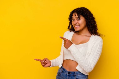 Sarı arka planda izole edilmiş genç melez kadın işaret parmağıyla fotokopi alanını işaret ederek şok oldu..