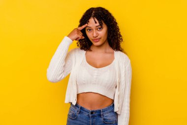 Sarı arka planda izole edilmiş genç melez bir kadın şakağı parmağıyla işaret ediyor, düşünüyor ve bir göreve odaklanıyor..