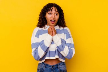 Genç melez kadın sarı arka planda izole edilmiş şans için dua ediyor, şaşırmış ve ağzı açık bir şekilde önüne bakıyor..