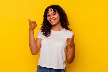 Sarı arka planda izole edilmiş genç bir ırk kadını. Baş parmağını kaldırıyor, gülümsüyor ve kendinden emin..