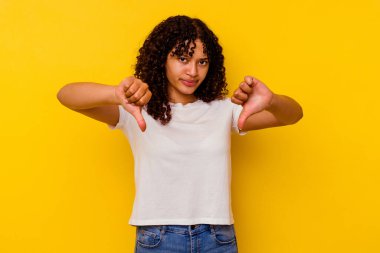 Sarı arka planda izole edilmiş genç melez kadın baş parmağını aşağı gösteriyor ve hoşlanmadığını ifade ediyor..