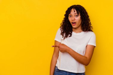 Sarı arka planda izole edilmiş genç melez kadın gülümsüyor ve boş alanda bir şeyler gösteriyor..