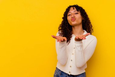 Sarı arka planda izole edilmiş genç melez bir kadın dudaklarını katlıyor ve hava öpücüğü yollamak için avuç içi tutuyor..