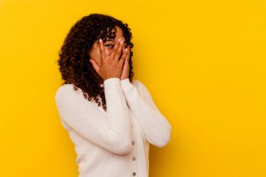 Sarı arka planda izole edilmiş genç bir ırk kadını parmaklarıyla kameraya göz kırpıyor ve yüzünü kapatmaktan utanıyor..