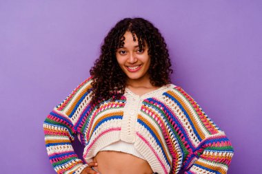 Mor arka planda izole edilmiş genç bir ırk kadını. Kendine güveniyor. Kalçaları tutuyor..