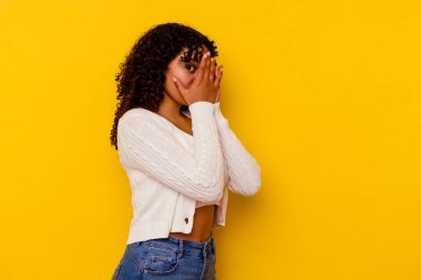 Sarı arka planda izole edilmiş genç melez kadın parmaklarının arasından korkmuş ve gergin bir şekilde göz kırp..