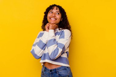 Sarı arka planda izole edilmiş genç melez kadın ellerini çenesinin altında tutuyor, mutlu bir şekilde yana bakıyor..