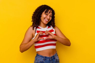 Sarı arka planda izole edilmiş genç melez kadın gülümsüyor ve elleriyle kalp şeklini gösteriyor..