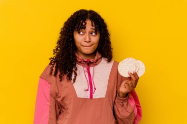 Sarı arka planda izole edilmiş pirinç keki tutan genç bir ırk sporcusu kafası karışmış ve kendinden emin değil..