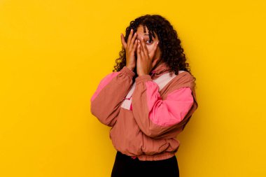 Pembe arka planda izole edilmiş genç melez kadın parmaklarının arasından korkmuş ve gergin bir şekilde göz kırpıyor..