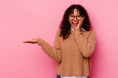 Pembe arka planda izole edilmiş genç melez kadın avuç içinde fotokopi alanı tutuyor, el üstünde tutuyor. Şaşırmış ve mutlu..