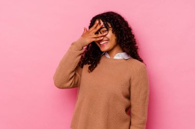 Genç melez kadın pembe arka planda izole edilmiş parmaklarıyla kameraya göz kırpıyor, yüzünü kapatmaktan utanıyor..