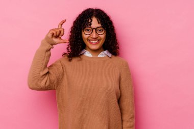 Pembe arka planda izole edilmiş genç melez bir kadın işaret parmaklarıyla küçük bir şey tutuyor, gülümsüyor ve kendinden emin..