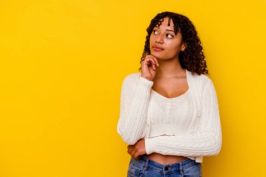 Sarı arka planda izole edilmiş genç melez kadın şüpheli ve şüpheci bir ifadeyle yana bakıyor..