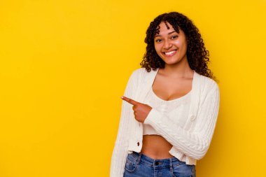 Sarı arka planda izole edilmiş genç melez kadın gülümsüyor ve boş alanda bir şeyler gösteriyor..