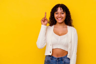 Sarı arka planda izole edilmiş genç melez kadın bir numarayı parmağıyla gösteriyor..