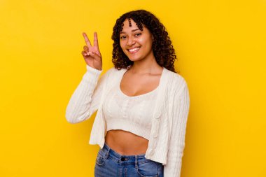 Sarı arka planda izole edilmiş genç melez kadın zafer işareti gösteriyor ve gülümsüyor..