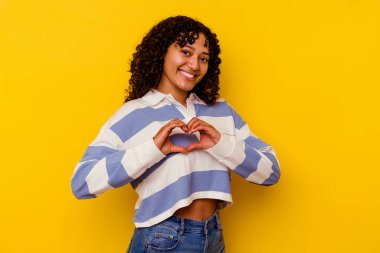 Sarı arka planda izole edilmiş genç melez kadın gülümsüyor ve elleriyle kalp şeklini gösteriyor..