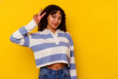 Sarı arka planda izole edilmiş genç melez kadın neşeli ve tasasız parmakları olan bir barış sembolü gösteriyor..