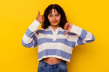 Sarı arka planda izole edilmiş genç, melez bir kadın baş parmağını kaldırıyor, zor seçim konsepti.