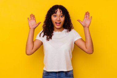 Sarı arka planda izole edilmiş genç melez kadın hoş bir sürpriz yaşıyor, heyecanlı ve el kaldırıyor..