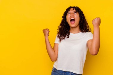 Genç melez kadın sarı arka planda izole edilmiş zafer sonrası yumruğunu kaldırıyor, kazanan konsept..