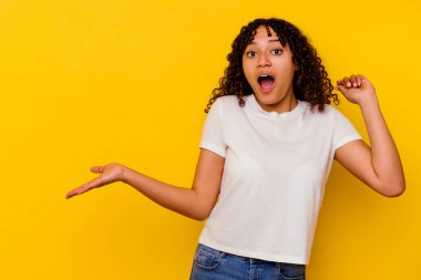 Sarı arka planda izole edilmiş genç melez kadın avuç içinde fotokopi alanı tutuyor, el üstünde tutuyor. Şaşırmış ve mutlu..