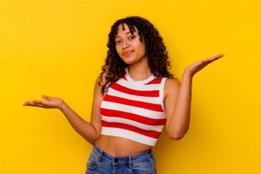 Sarı arka planda izole edilmiş genç melez bir kadın. Kafası karışmış ve omuz silkiyor. Kopya alanını tutmak için..