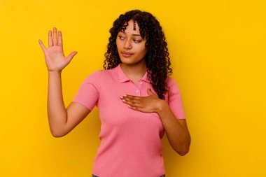 Genç melez kadın sarı arka planda izole edilmiş yemin ediyor, elini göğsüne koyuyor..