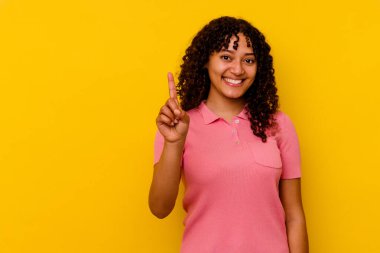 Sarı arka planda izole edilmiş genç melez kadın bir numarayı parmağıyla gösteriyor..