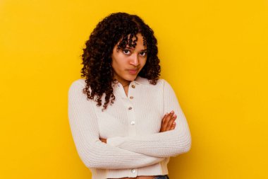 Sarı arka planda izole edilmiş, hoşnutsuzca kaşlarını çatan, kollarını kavuşturan genç melez bir kadın..