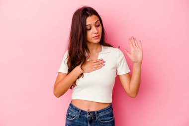 Pembe arka planda izole edilmiş genç Hintli kadın yemin ediyor, elini göğsüne koyuyor..