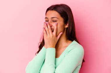 Pembe arka planda izole edilmiş genç Hintli bir kadın bir şeye gülüyor, elleriyle ağzını kapatıyor..
