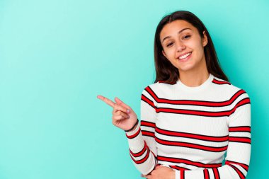 Mavi arka planda izole edilmiş genç Hintli kadın işaret parmağıyla işaret ederek gülümsüyor..
