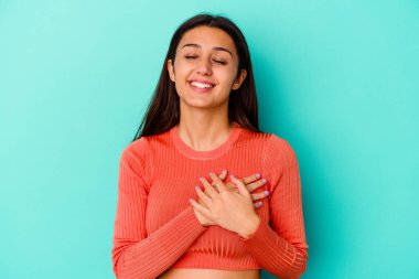 Mavi arka planda izole edilmiş genç Hintli kadın gülüyor ellerini kalbe koyuyor, mutluluk kavramını koruyor..