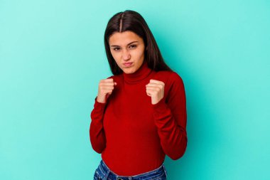 Mavi arka planda izole edilmiş genç Hintli kadın kameraya yumruk gösteriyor, agresif yüz ifadesi..