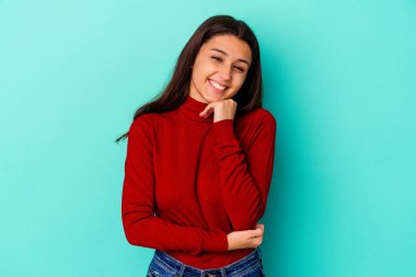 Genç Hintli bir kadın mavi arka planda izole edilmiş mutlu ve kendinden emin gülümsüyor, elleriyle çenesine dokunuyor..